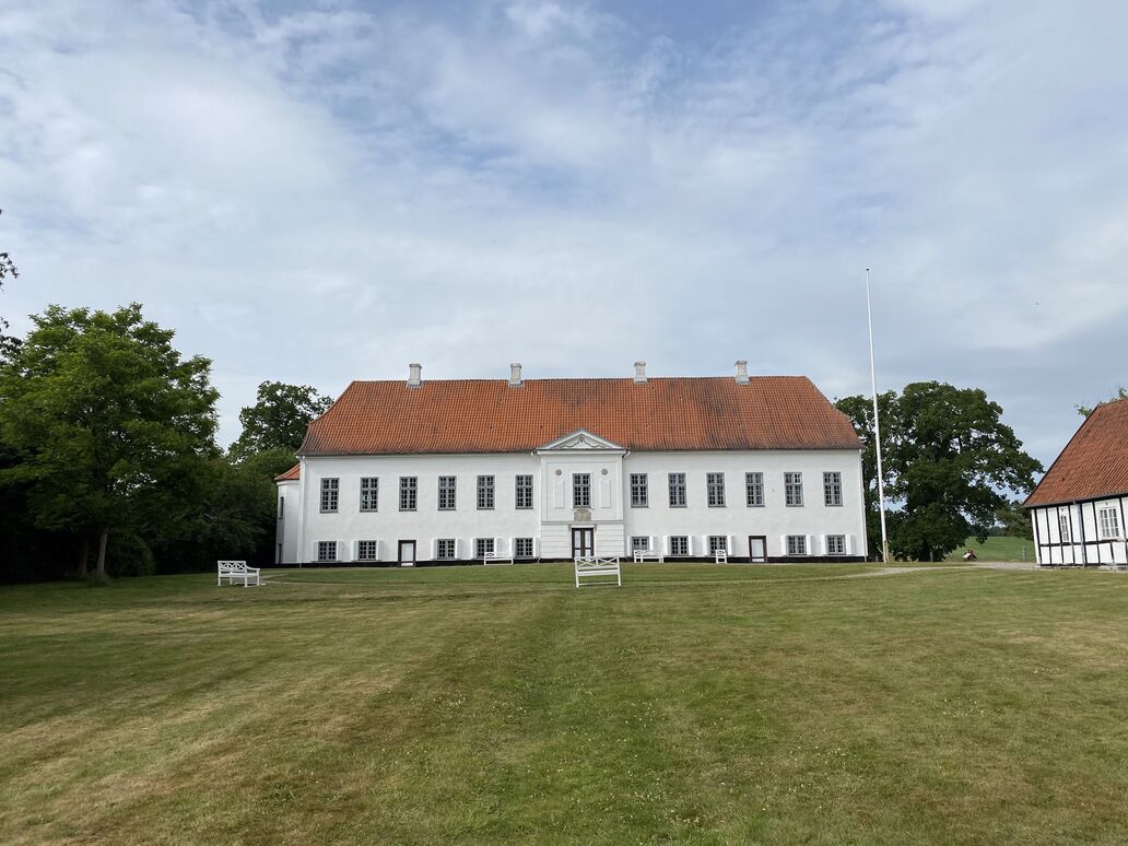 Herregården Fussingøs hovedbygning set fra havesiden. Hvidkalket hovedbygning i klassicistisk stil. Dele af hvid bindingsværksbygning ses til højre i billedet.