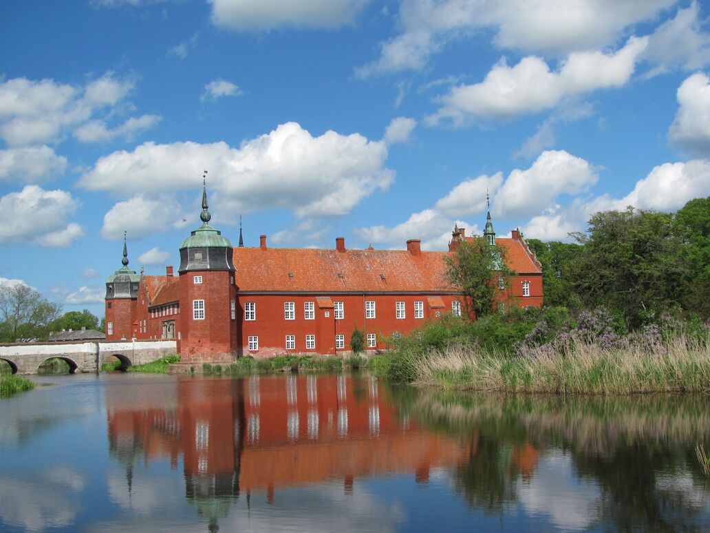 Herregården Løvenborg med voldgrav i forgrunden. Rødkalket hovedbygning med hjørnetårne med spir, mens renæssancegavle anes mod højre i billedet. Til venstre ses bro over voldgrav.